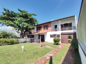 Sobrado em frente ao mar com grande jardim verde, área coberta com cadeiras e sacadas com vista ao mar, e árvore com sombra.