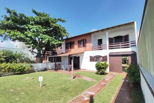 Sobrado em frente ao mar com grande jardim verde, área coberta com cadeiras e sacadas com vista ao mar, e árvore com sombra.